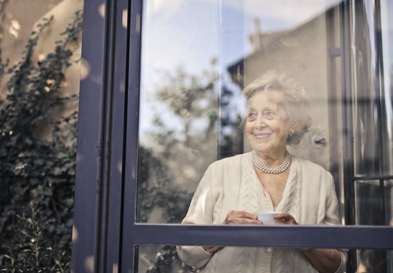 Femme souriante regardant par la fenêtre, tasse en main.