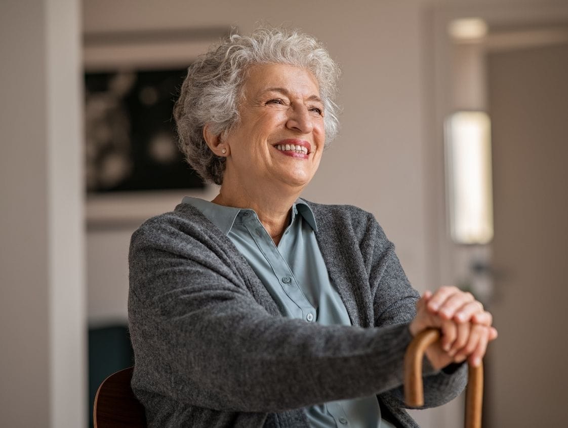 Femme âgée souriante avec canne assise chez elle.