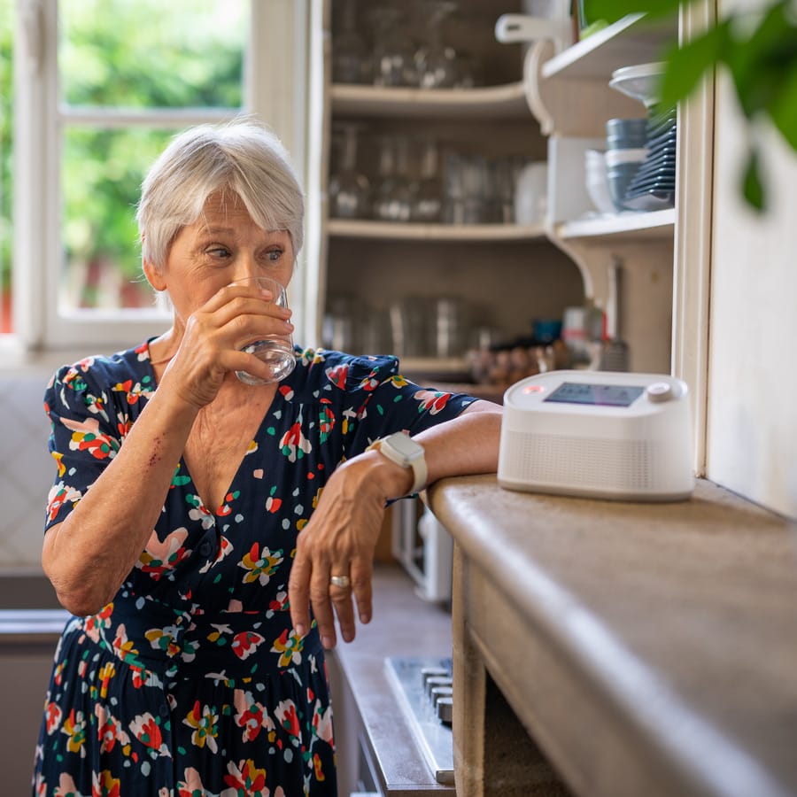 Femme âgée buvant de l'eau dans cuisine lumineuse.